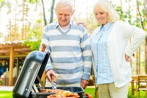 njuter grilla tillsammans. Lycklig senior par grilla kött på de grill medan stående på de tillbaka gård av deras hus foto