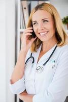 ger råd på de telefon. Lycklig kvinna läkare i vit enhetlig talande på de mobil telefon och leende foto