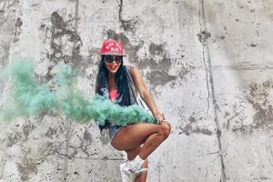 färg de värld. lekfull ung kvinnor innehav rök bomba och leende medan Framställ mot de betong vägg foto