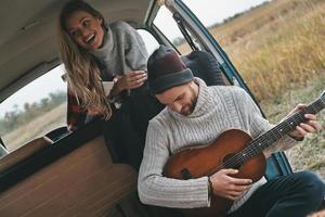 njuter Lycklig ögonblick. stilig ung man spelar gitarr för hans skön flickvän medan Sammanträde i retro stil mini skåpbil foto