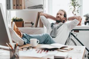 njuter liten ha sönder. stilig ung man med stängd ögon innehav händer Bakom huvud och vilar med fötter på skrivbord medan Sammanträde på arbetssätt plats i kreativ kontor foto