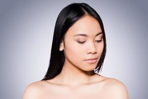 ren skönhet. porträtt av skön ung och bar överkropp asiatisk kvinna ser ner medan stående mot grå bakgrund foto