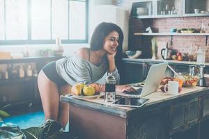surfing webb från kök. attraktiv ung afrikansk kvinna lutande på de kök skrivbord och ser på bärbar dator med leende foto
