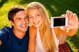 fångst en ljus ögonblick. Lycklig ung kärleksfull par framställning selfie och leende medan Sammanträde tillsammans på de gräs i parkera foto
