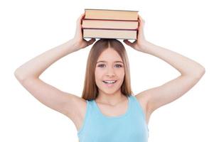 smart skolflicka. glad Tonårs flicka innehav bok stack på henne huvud och leende medan stående isolerat på vit foto