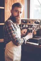 Barista på arbete. ung skäggig man i förkläde framställning kaffe och ser på kamera medan stående på Kafé foto