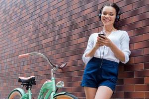 sorglös tid. skön ung kvinna i hörlurar lyssnande till mp3 spelare och leende medan stående nära henne årgång cykel foto