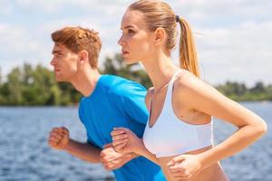 sport är vår liv. glad ung kvinna och man i sporter Kläder löpning längs de flodbank foto