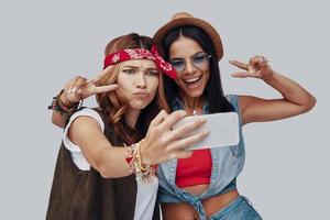 två attraktiv eleganta ung kvinnor tar selfie och leende medan stående mot grå bakgrund foto