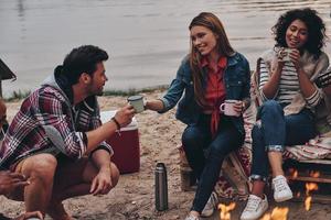 njuter värma te. grupp av ung människor i tillfällig ha på sig leende och innehav muggar medan camping nära de bål foto
