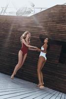 spontan dansa. full längd av två attraktiv ung kvinnor i badkläder leende medan dans mot de trä- vägg utomhus foto