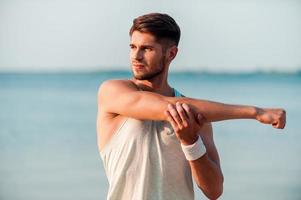 man avmaskning upp. självsäker ung muskulös man stretching hans vapen medan stående på de havet foto