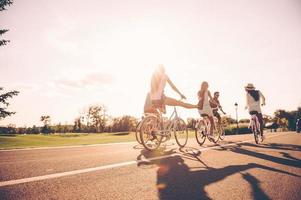 njuter trevlig sommar dag tillsammans. låg vinkel se av ung människor ridning cyklar längs en väg och ser Lycklig foto