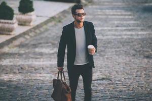 eleganta stilig på de gå. stilig ung man i smart tillfällig ha på sig bärande väska och kaffe kopp medan gående längs de gata foto