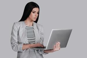 arbetssätt på henne ny bärbar dator. skön ung kvinna i smart tillfällig ha på sig arbetssätt på bärbar dator medan stående mot grå bakgrund foto