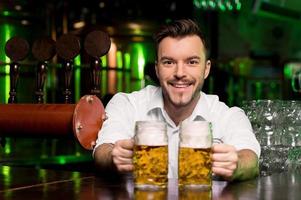 nyligen knackade öl. stilig smiley bartender stretching ut öl muggar och leende foto