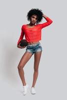 redo till spela. full längd studio skott av attraktiv ung afrikansk kvinna i tillfällig ha på sig förvaring hand i hår och bärande en boll medan stående mot grå bakgrund foto