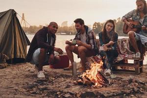 Allt de behöver är varje Övrig. grupp av ung människor i tillfällig ha på sig leende medan njuter strand fest nära de lägereld foto