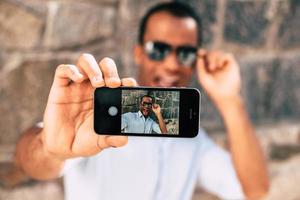 selfie roligt. stilig ung afrikansk man framställning selfie med hans smart telefon och ser Lycklig medan stående mot de urkärnad vägg utomhus foto