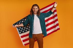 Lycklig ung man i tillfällig Kläder ser på kamera och leende medan innehav amerikan flagga mot gul bakgrund foto