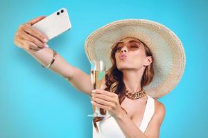skön ung leende kvinna i elegant hatt innehav champagne flöjt och framställning selfie foto