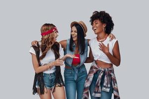 tre attraktiv eleganta ung kvinnor ser på varje Övrig och skrattande medan stående mot grå bakgrund foto