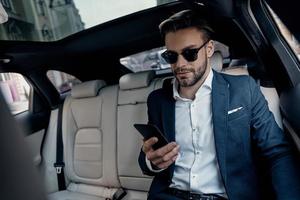 organisering varje minut. stilig ung man i full kostym ser på hans smart telefon medan Sammanträde i de bil foto