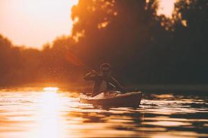 Kajakpaddling är hans livsstil. ung man Kajakpaddling på flod med solnedgång i de bakgrund foto