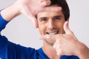 fokusera på du. lekfull ung man i blå Tröja gestikulerar finger ram och leende medan stående mot grå bakgrund foto
