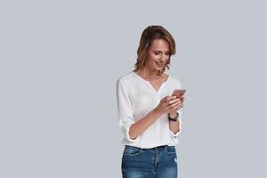 tid till ge ett svar. attraktiv ung kvinna i tillfällig ha på sig använder sig av henne smart telefon och leende medan stående mot grå bakgrund foto