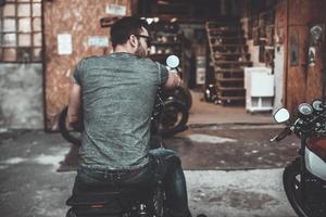 verklig cyklist. bak- se av självsäker ung man Sammanträde på hans cykel med motorcykel garage i de bakgrund foto