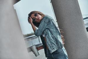 alltid i stil. attraktiv ung kvinna i jeans ha på sig innehav en disponibel kopp och justeras henne hår medan stående nära de arkitektonisk kolumn utomhus foto