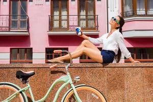 njuter henne fri tid i stad. skön ung leende kvinna i skraj solglasögon Sammanträde utomhus och nära henne årgång cykel foto
