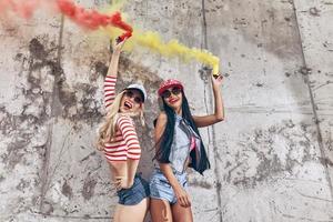 färgrik roligt. två Lycklig ung kvinnor innehav rök bomber och leende medan Framställ mot de betong vägg foto