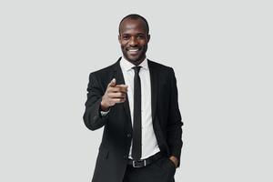 stilig ung afrikansk man i formell klädsel pekande du och leende medan stående mot grå bakgrund foto