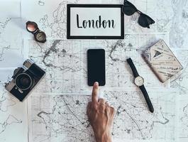 London är ny destination. stänga upp topp se av man använder sig av smart telefon med solglasögon, Foto kamera, kompass, Kolla på och pass liggande på Karta runt om
