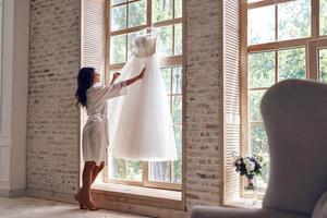 utseende bara höger. full längd av skön ung kvinna i silke morgonrock rörande henne bröllop klänning medan stående nära de fönster foto