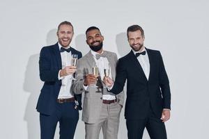 tre stilig ung män i kostymer och flugor toasting med champagne medan stående mot grå bakgrund foto