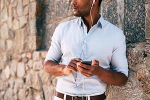 man njuter musik. närbild av stilig ung afrikansk man i hörlurar bärande smart telefon medan stående mot de urkärnad vägg utomhus foto