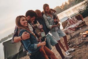 Lycklig till vara runt om. grupp av ung människor i tillfällig ha på sig leende och fattande medan njuter strand fest nära de sjö foto