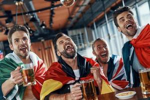 Lycklig ung män täckt i internationell flaggor dricka öl och tittar på sport spel medan Sammanträde i de pub foto