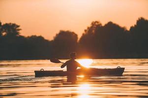 njuter de bäst solnedgång på flod. sida se av ung man Kajakpaddling på flod med solnedgång i de bakgrund foto