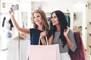 selfie efter framgångsrik handla. två skön kvinnor med handla påsar framställning selfie medan stående på de Kläder Lagra foto