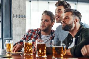 tre ung män i tillfällig Kläder dricka öl och tittar på sport spel medan Sammanträde i de pub foto