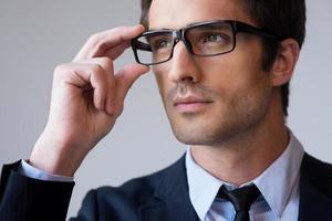 självsäker och framgångsrik. porträtt av självsäker ung man i formell klädsel justeras hans glasögon och ser bort medan stående mot grå bakgrund foto