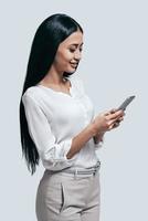 skriver ett Viktig företag meddelande. ung självsäker kvinna i tillfällig ha på sig innehav smart telefon och ser på skärm med en leende på henne ansikte medan stående mot grå bakgrund foto