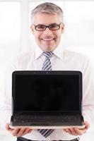 din annons här. leende senior man i skjorta och slips innehav bärbar dator och som visar de övervaka till du foto
