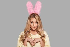 sötare än några kanin. attraktiv ung kvinna i rosa kanin öron gestikulerar och ser på kamera medan stående mot grå bakgrund foto