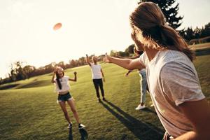 grupp av ung människor i tillfällig ha på sig spelar frisbee medan utgifterna sorglös tid utomhus foto