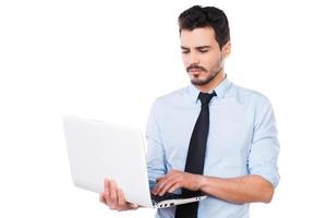 självsäker företag expert. stilig ung man i skjorta och slips arbetssätt på bärbar dator medan stående mot vit bakgrund foto